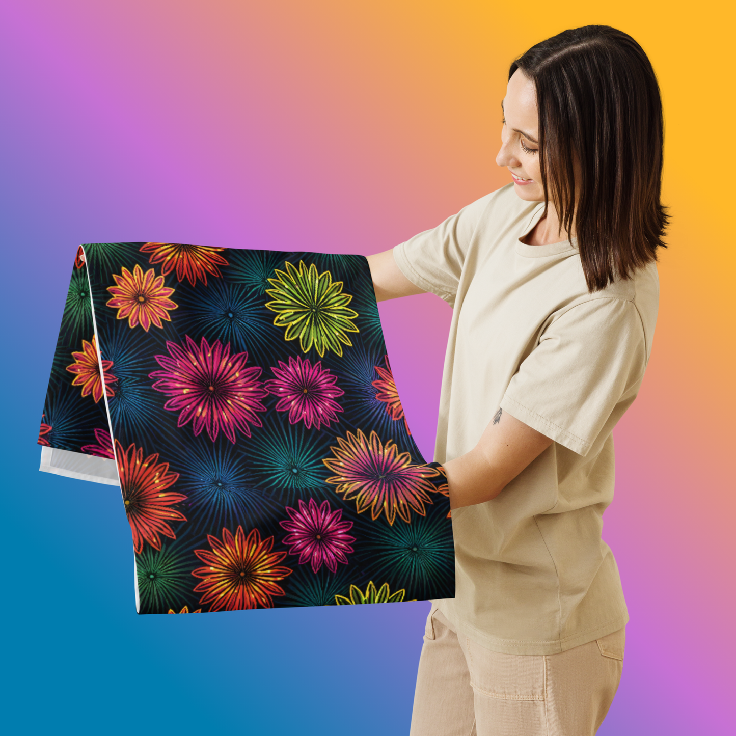 Fluorescent Floral Table Runner hero lifestyle shot — smiling White female model in cream oversized tee holding folded table runner showing all-over print of neon pink orange yellow-green and teal daisy-like flowers with radiating petal details on dark navy background against blue to orange gradient background