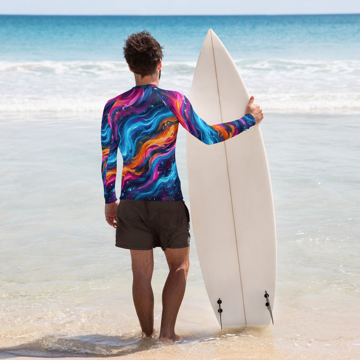 Surfer holding a white surfboard on a tropical beach wearing the Cosmic Surge men's rash guard, back view showing swirling neon cosmic wave print in blue, pink, and orange