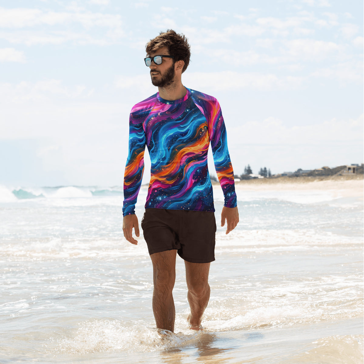 Man walking along the beach shoreline wearing the Cosmic Surge neon galaxy rash guard with sunglasses and black swim shorts, ocean waves in the background