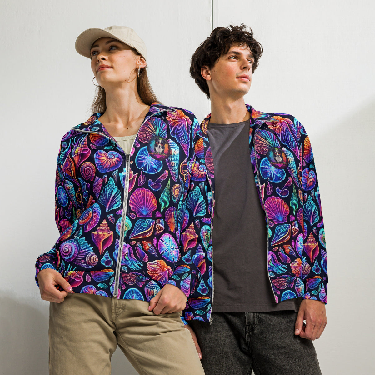 Female and male models wearing the Chromashell Unisex Track Jacket side by side, showing the vibrant neon seashell all-over print in purple, pink, teal, and orange on black