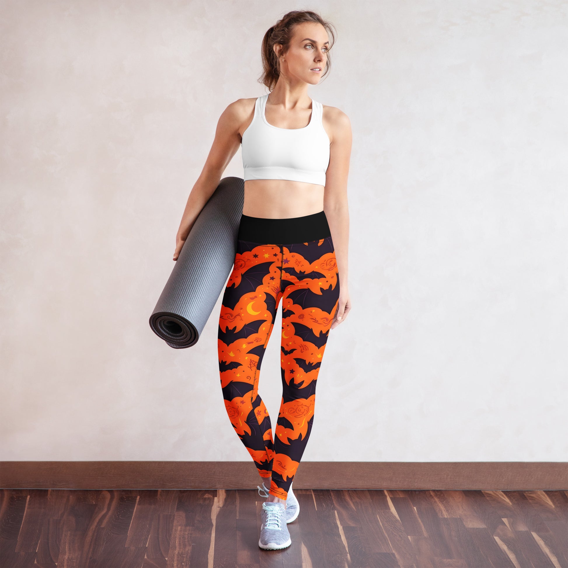 Fright Night Yoga Leggings lifestyle shot — White female model in white sports bra carrying yoga mat wearing leggings showing all-over orange bat and dark navy cloud Halloween print with black high-waist band on neutral studio background with wood floor