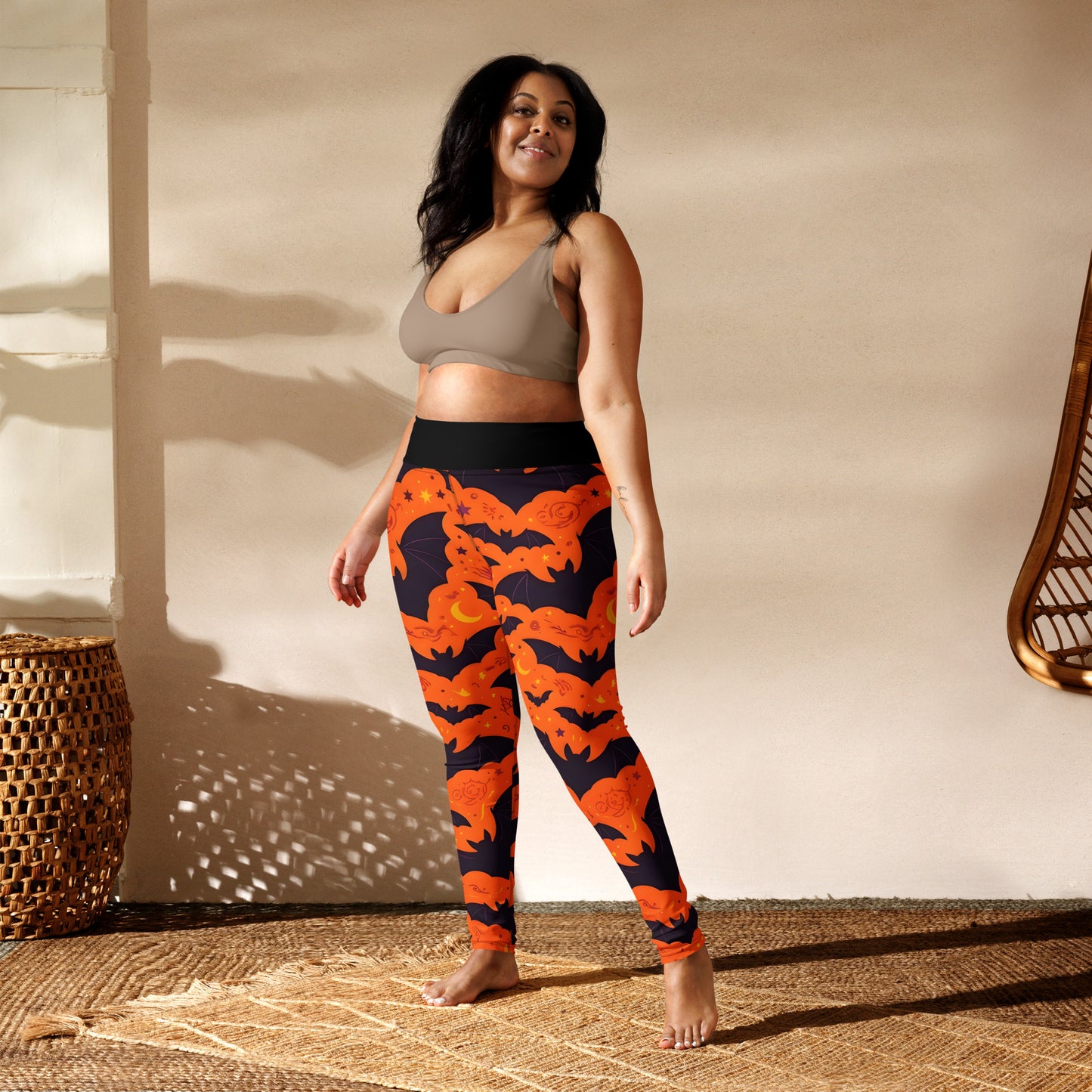 Fright Night Yoga Leggings lifestyle shot — smiling plus-size Latina female model in taupe sports bra wearing leggings showing all-over orange bat and dark navy cloud Halloween print with black high-waist band in boho studio with rattan furniture and woven rug in warm sunlight