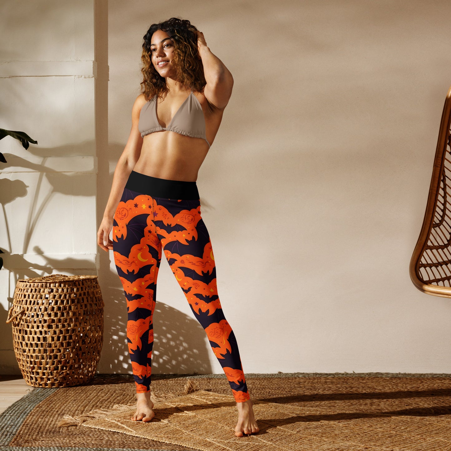 Fright Night Yoga Leggings lifestyle shot — smiling Latina female model in taupe triangle bralette wearing leggings showing all-over orange bat and dark navy cloud Halloween print with black high-waist band in boho studio with rattan furniture and woven rug in warm sunlight