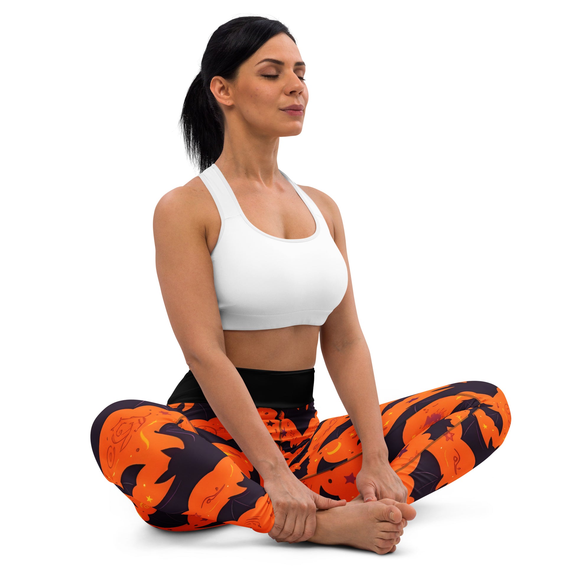 Fright Night Yoga Leggings lifestyle shot — White female model in white sports bra seated in cross-legged yoga pose wearing leggings showing all-over orange bat and dark navy cloud Halloween print with black high-waist band on white background