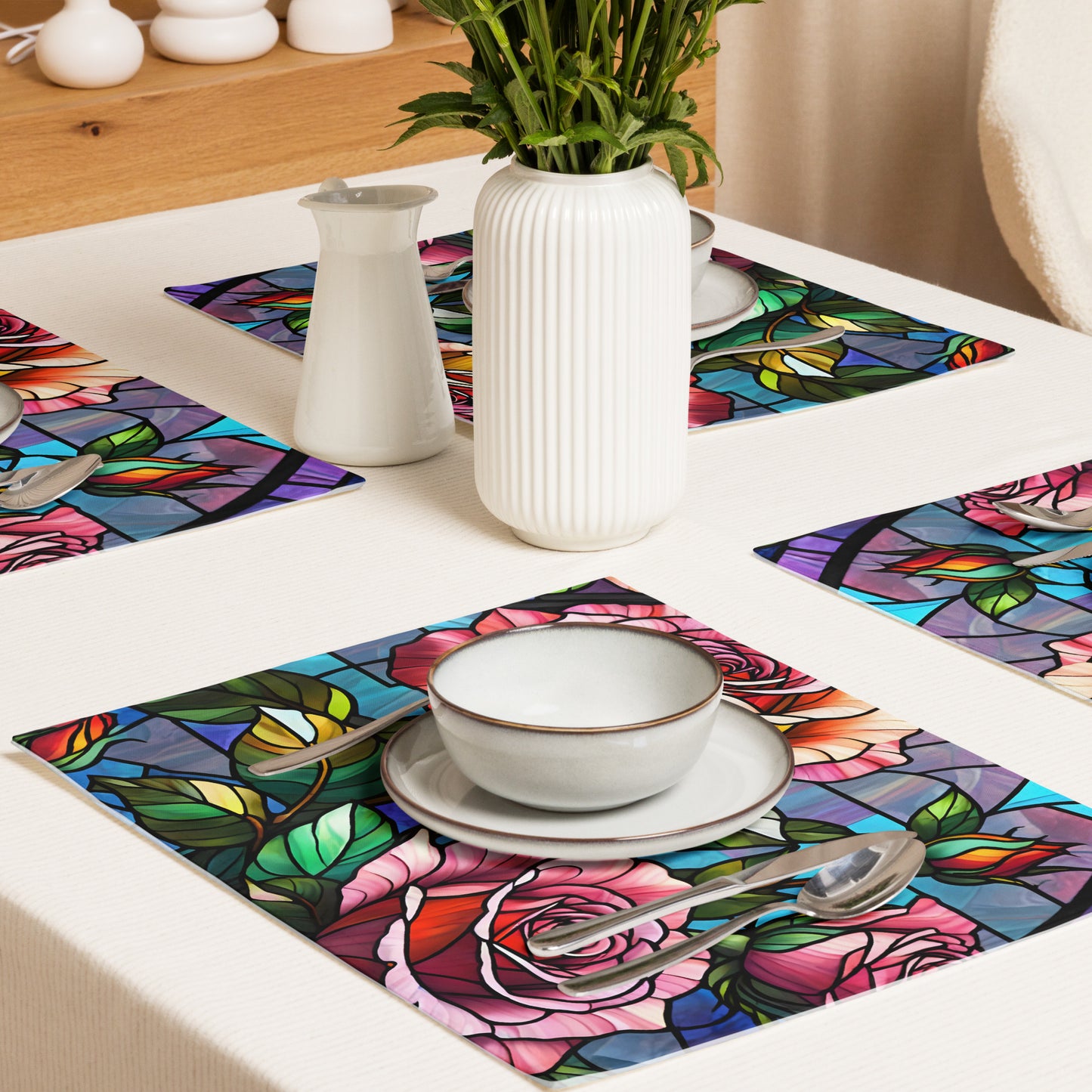 Stained Roses Placemat Set product shot — single placemat flat lay with second placemat folded beneath showing stained glass mosaic design of large pink and red roses with green leaves and colorful geometric background panels in blue purple and teal with white plate fork and spoon on white background