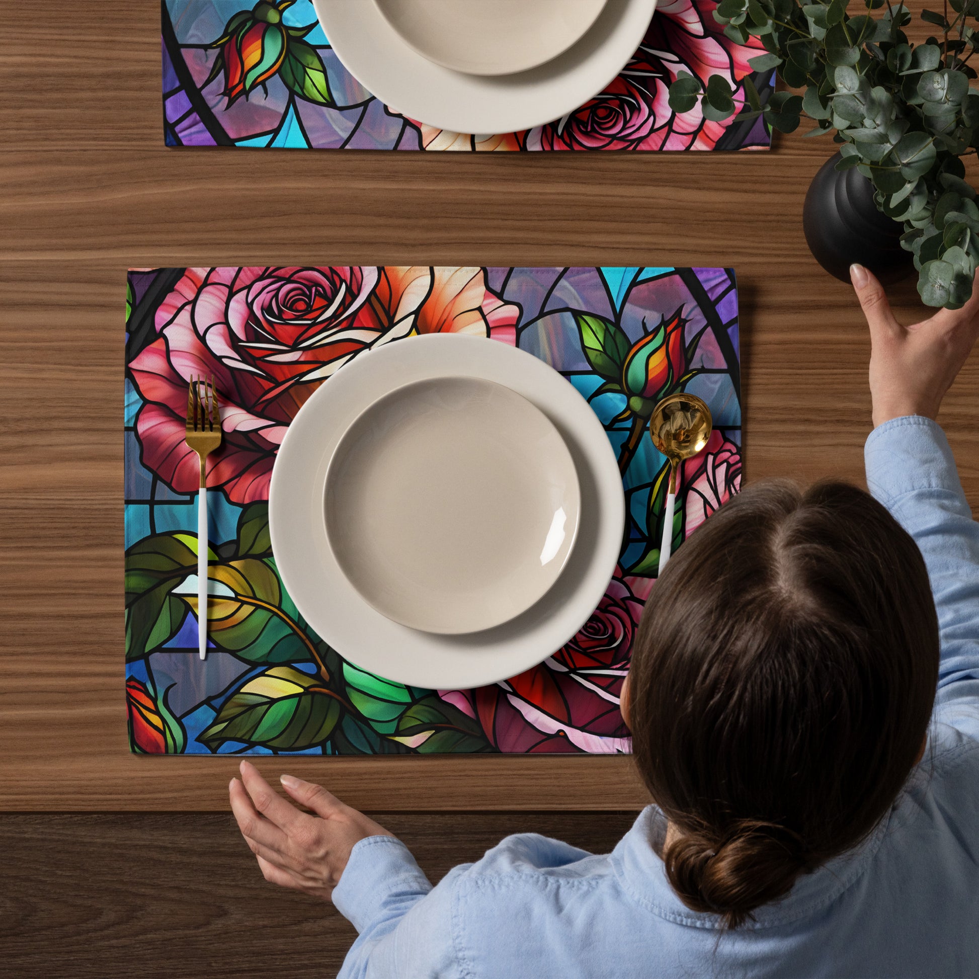 Stained Roses Placemat Set lifestyle shot — overhead view of White female model in light blue shirt placing white plate on placemat on dark wood dining table with gold cutlery and second placemat and dark vase of eucalyptus in background showing stained glass mosaic design of large pink and red roses with green leaves