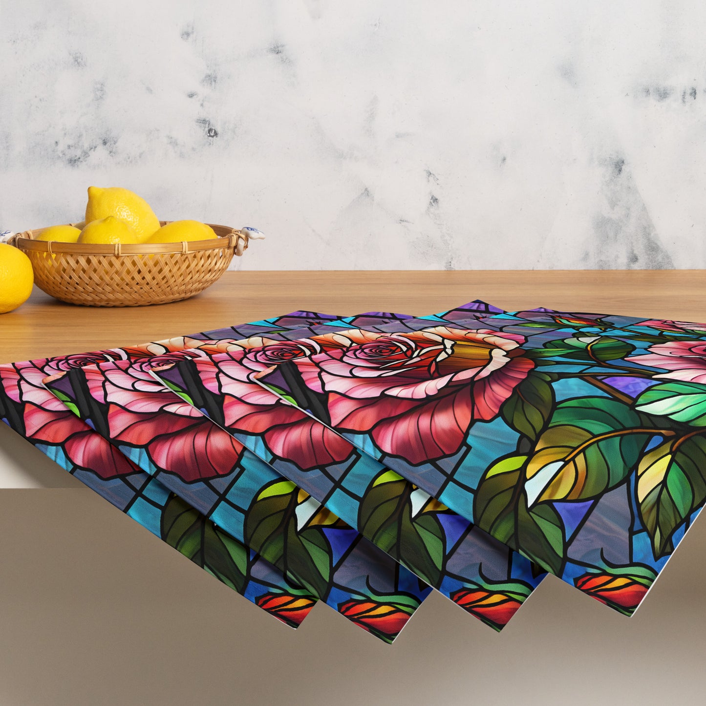 Stained Roses Placemat Set lifestyle shot — set of four placemats stacked and fanned on wood countertop beside wicker basket of lemons against marble wall showing stained glass mosaic design of large pink and red roses with green leaves and colorful geometric background panels in blue purple and teal