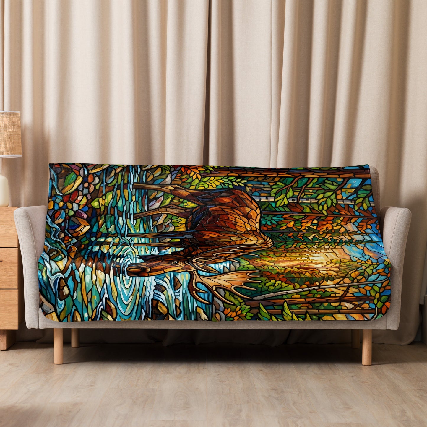 Mosaic Moose Sherpa Blanket lifestyle shot — 50x60 blanket draped over cream sofa showing full stained glass mosaic design of bull moose drinking from rocky mountain stream with autumn forest canopy in orange green and gold against neutral curtain background with wood nightstand and lamp