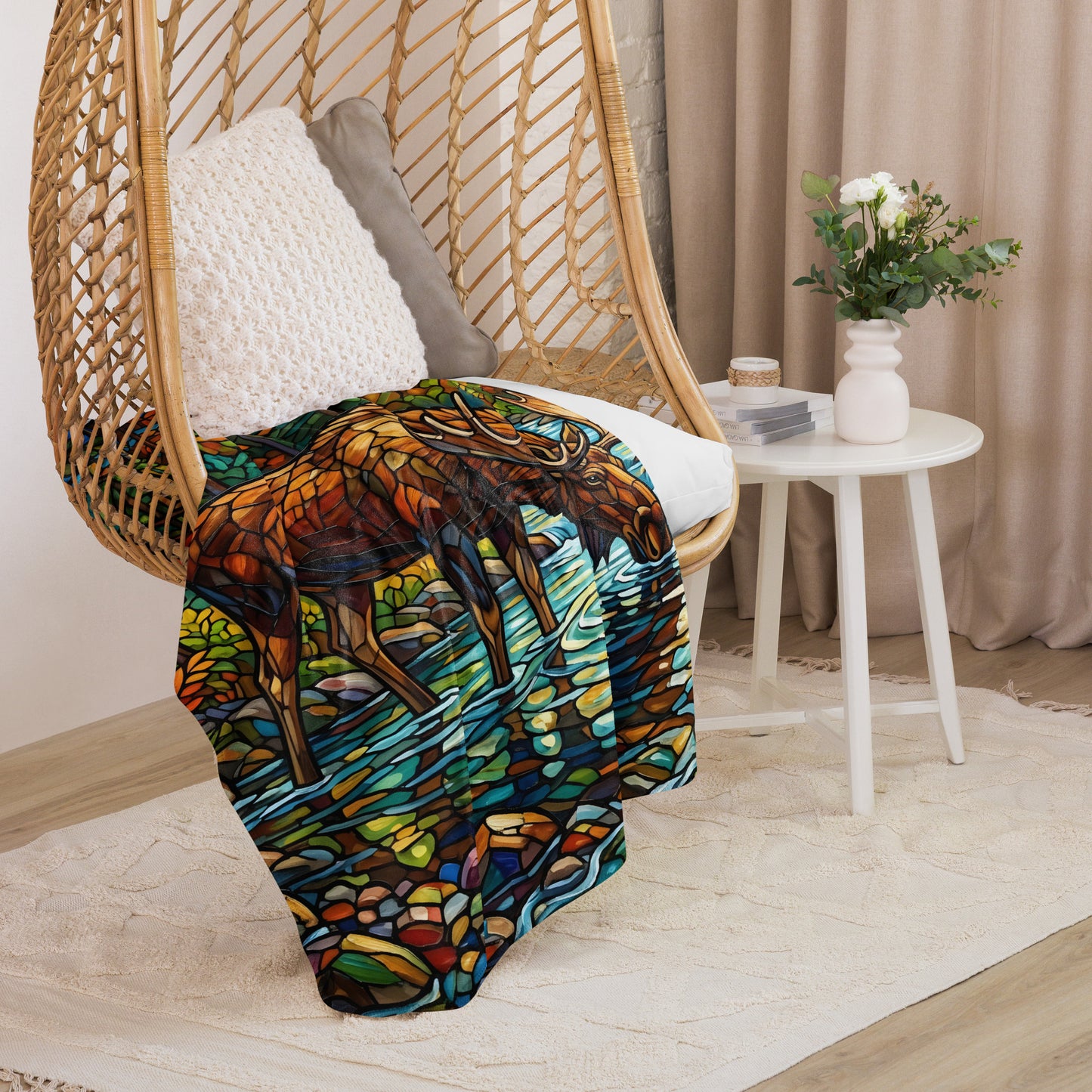 Mosaic Moose Sherpa Blanket lifestyle shot — 60x80 blanket draped over rattan egg chair showing stained glass mosaic bull moose drinking from stream with autumn forest detail and cream sherpa interior visible with white side table flowers against neutral curtain background