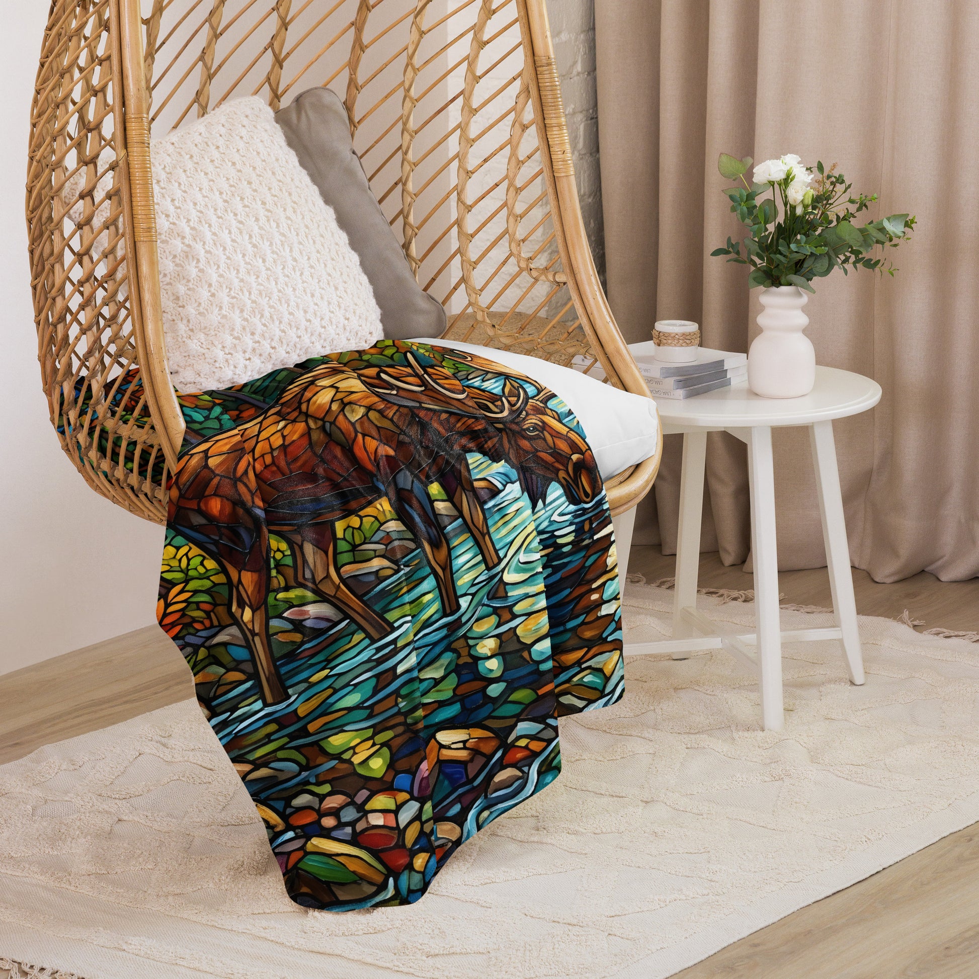 Mosaic Moose Sherpa Blanket lifestyle shot — 60x80 blanket draped over rattan egg chair showing stained glass mosaic bull moose drinking from stream with autumn forest detail and cream sherpa interior visible with white side table flowers against neutral curtain background