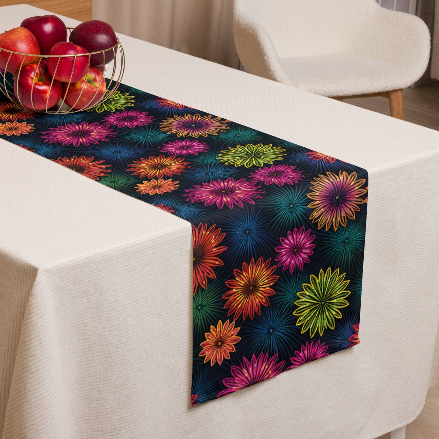 Fluorescent Floral Table Runner lifestyle shot — table runner laid across white dining table with gold wire fruit bowl of red apples and cream dining chair in background showing all-over print of neon pink orange yellow-green and teal daisy-like flowers with radiating petal details on dark navy background