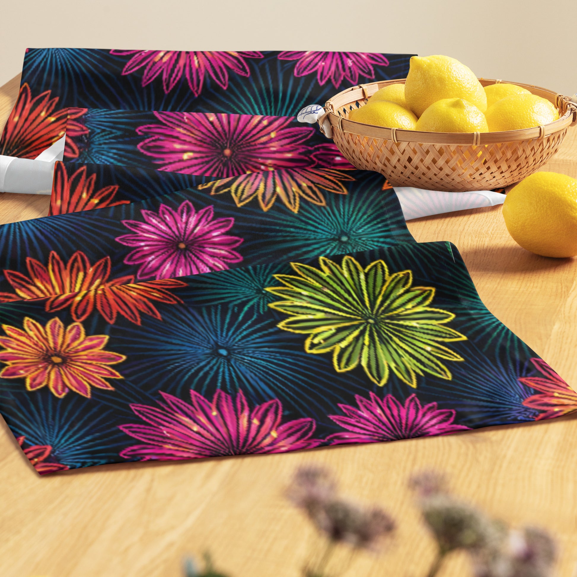 Fluorescent Floral Table Runner lifestyle shot — table runner laid flat on light wood dining table beside wicker basket of lemons showing close-up of neon pink orange yellow-green and teal daisy-like flowers with radiating petal details on dark navy background