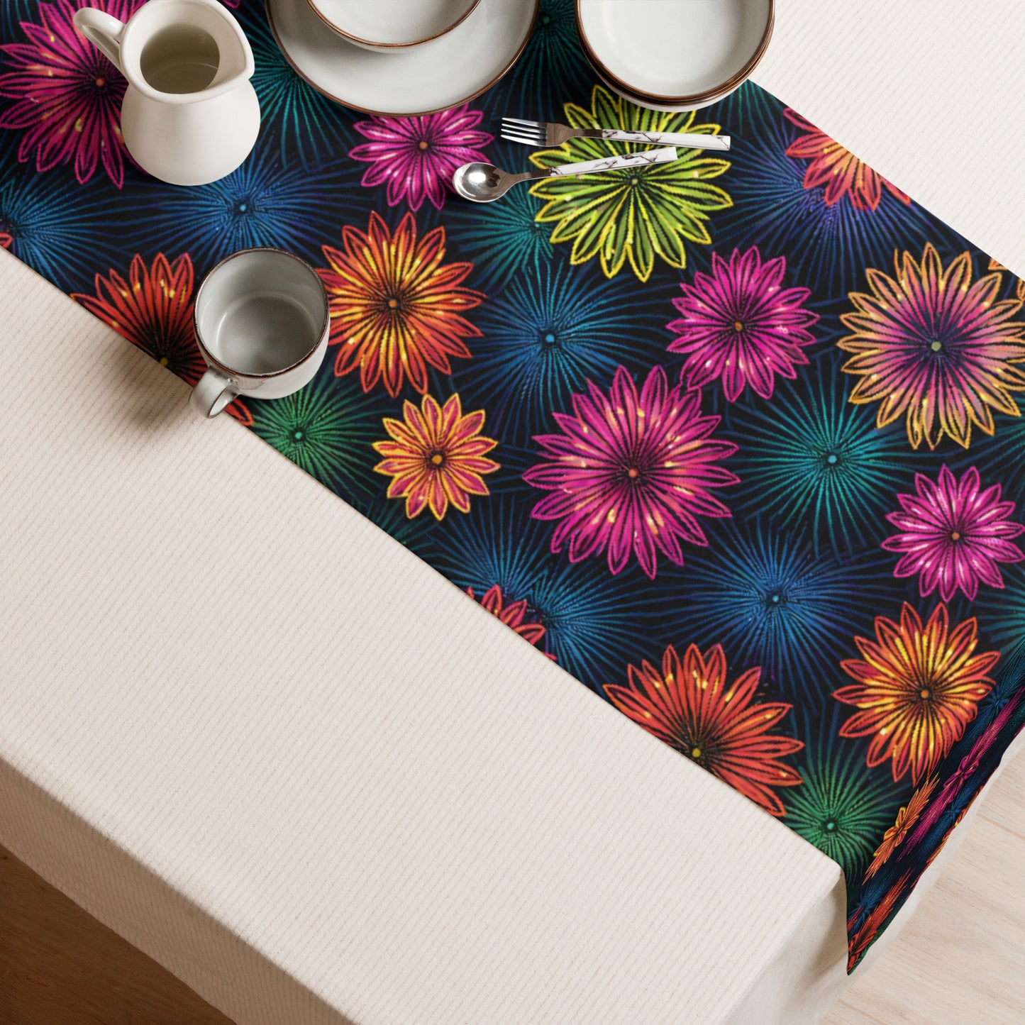 Fluorescent Floral Table Runner lifestyle shot — overhead view of table runner on white linen tablecloth set with white plates cups and silver cutlery showing all-over print of neon pink orange yellow-green and teal daisy-like flowers with radiating petal details on dark navy background